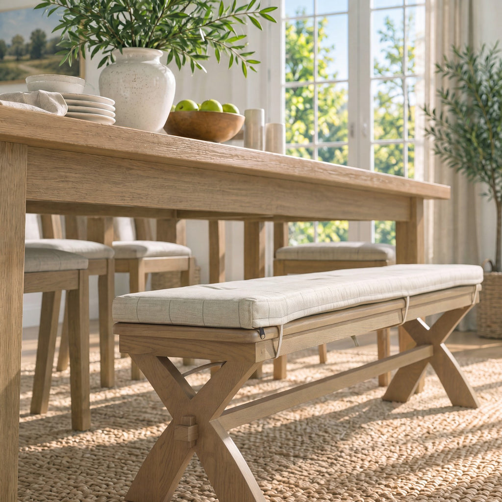 A light natural wood dining bench with a thick cream upholstered cushion sits beneath a matching wooden dining table in a bright, sunlit dining room.