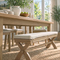 A light natural wood dining bench with a thick cream upholstered cushion sits beneath a matching wooden dining table in a bright, sunlit dining room.