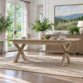 A light natural wood dining table with an X-shaped base and a matching sideboard in a bright, modern dining room. The table is styled with green plants in ceramic vases, candles, and a bowl of fruit, set on a textured beige rug.