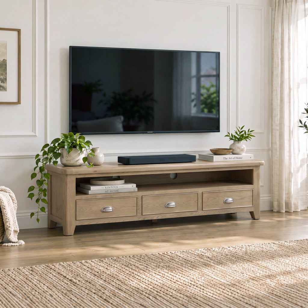 A light natural wood TV stand with three drawers and silver handles, styled in a bright living room under a wall-mounted television.