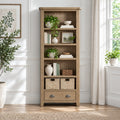 A tall, rustic five-shelf wooden bookcase with a single drawer at the bottom, styled with books, potted plants, and wicker baskets in a bright, modern room.