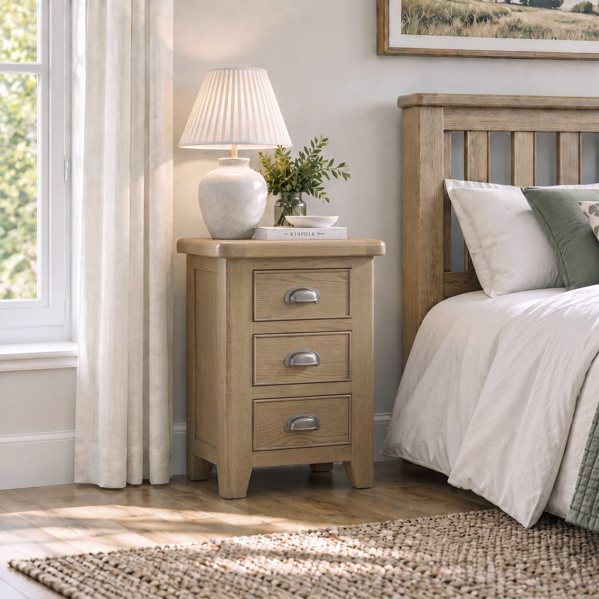 A light natural wood three-drawer nightstand with silver metal handles, styled next to a bed with white linens and a wooden headboard in a bright bedroom.