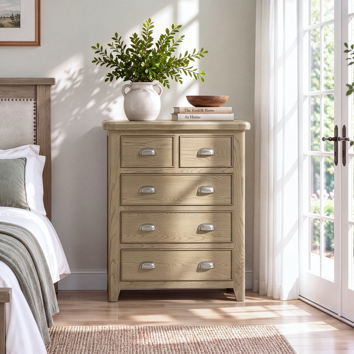 A light oak wood five-drawer nightstand or dresser with silver metal handles, styled in a bright bedroom next to a bed and a potted plant.