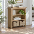 A light oak wood three-tier bookcase with two woven storage baskets on the bottom shelf, styled with books, potted plants, and a vase in a bright room.