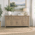 A light beige wood sideboard or console cabinet with three drawers and three cabinet doors, styled in a bright living room setting with a vase of flowers, a lamp, and a framed landscape painting on the wall.