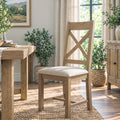 A light natural wood dining chair with a cushioned seat and crossed backrest slats, shown in a bright dining room setting next to a wooden table.