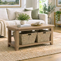 A light oak wood coffee table with two woven storage baskets underneath, styled in a bright, modern living room next to a white sofa.