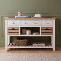A white and light wood console table styled in a room setting with a green paneled wall, wooden floor, and a beige rug. The table features three white drawers on top, open shelving with wooden crate-style fronts, and a lower slatted shelf holding a wicker basket and books.