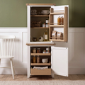 A tall white and light oak kitchen cabinet with two doors open, showing shelves and a drawer stocked with jars, bottles, and packaged goods. The cabinet stands in a kitchen setting with a green wall, white wainscoting, a wooden floor, and a white chair partially visible on the left.