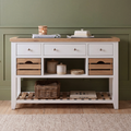 A white and light wood console table styled in a room setting with a green paneled wall, wooden floor, and a beige rug. The table features three white drawers on top, open shelving with wooden crate-style fronts, and a lower slatted shelf holding a wicker basket and books.