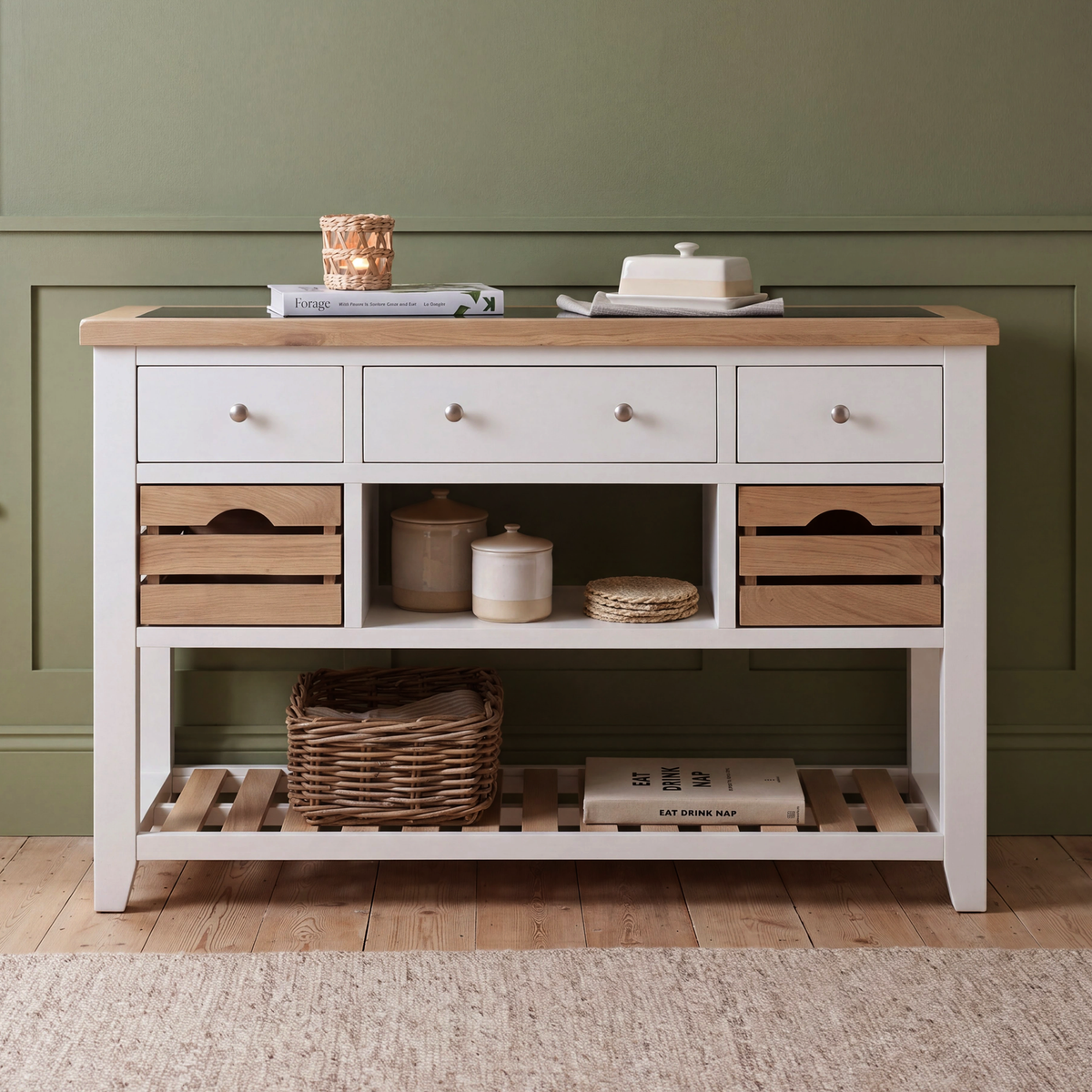A white and light wood console table styled in a room setting with a green paneled wall, wooden floor, and a beige rug. The table features three white drawers on top, open shelving with wooden crate-style fronts, and a lower slatted shelf holding a wicker basket and books.