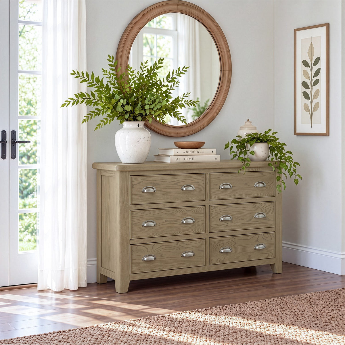 A light sage green wooden six-drawer dresser styled in a bright room with a round wooden mirror, white vases with green plants, and books on top.