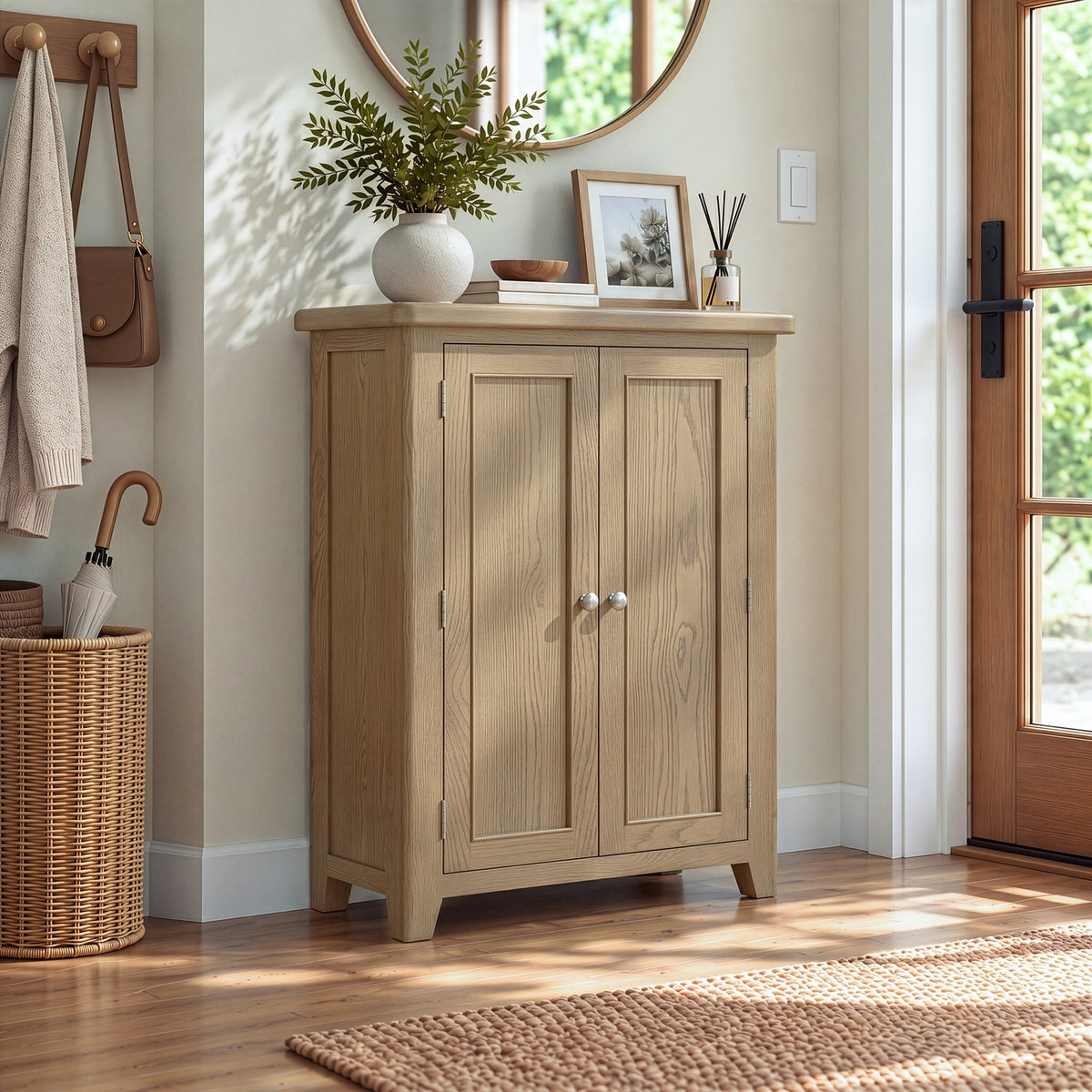 A light oak wood entryway cabinet with two doors and silver knobs, styled in a bright entryway with a plant, picture frame, and a wicker basket nearby.