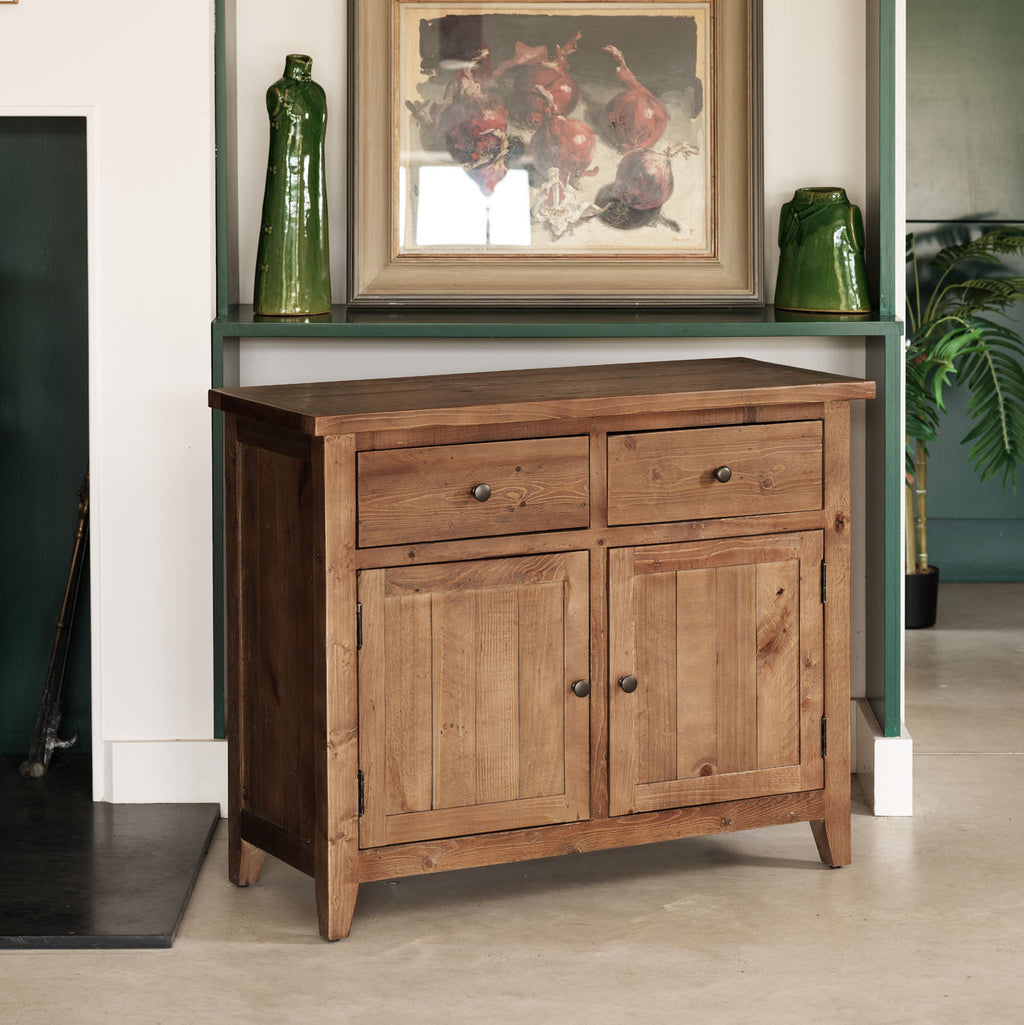 A rustic wooden sideboard cabinet with two drawers on top and two paneled doors below, shown in a styled interior setting with a green mantel, framed artwork, and decorative vases.