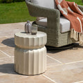 A small round outdoor side table with a fluted pedestal design in a warm beige finish, styled on a stone patio next to a wicker patio chair with a rust-colored throw blanket.