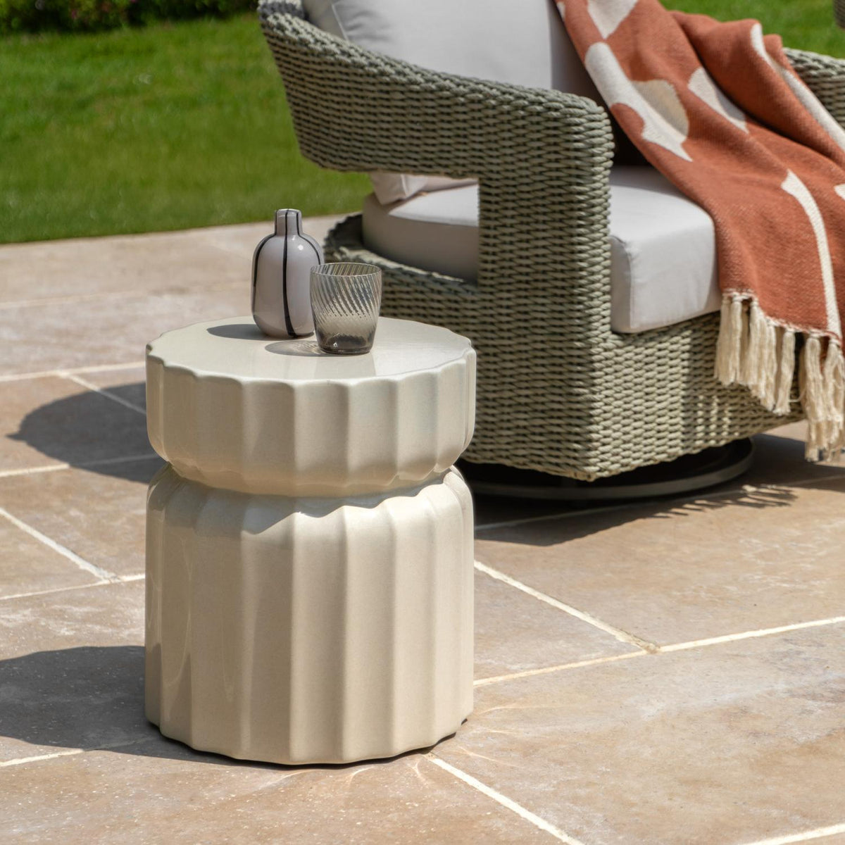 A small round outdoor side table with a fluted pedestal design in a warm beige finish, styled on a stone patio next to a wicker patio chair with a rust-colored throw blanket.