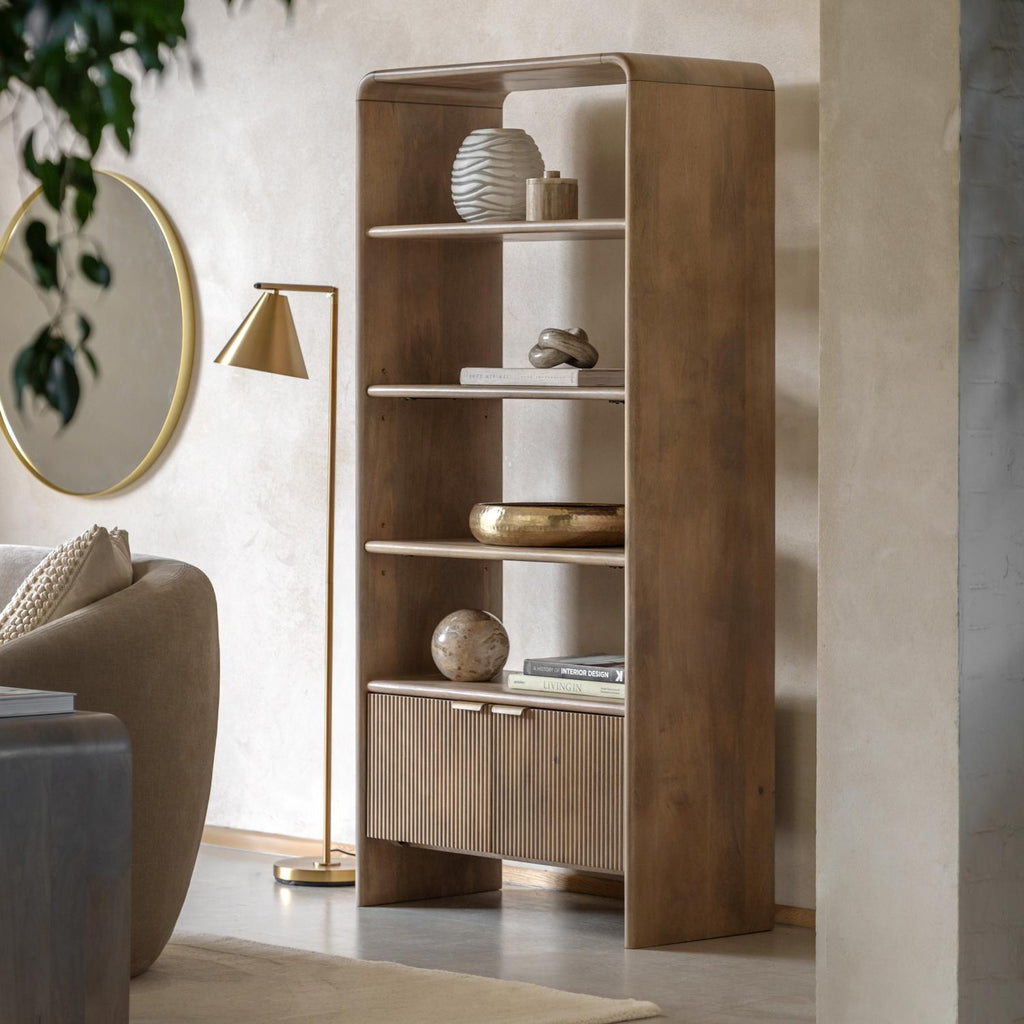 A tall wooden bookcase with open shelves and two slatted cabinet doors stands in a styled living room. The shelves hold decorative vases, books, and small objects, and the room includes a floor lamp, round mirror, sofa, and rug.