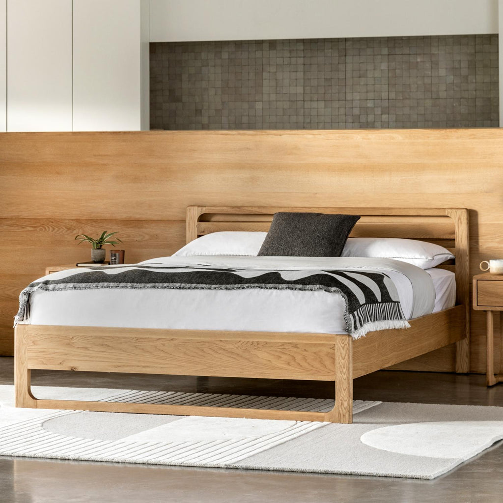 A modern light oak wood bed frame with a low-profile footboard and headboard, styled with white bedding, a black throw pillow, and a black-and-white striped blanket, set in a minimalist bedroom.