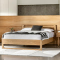 A modern light oak wood bed frame with a low-profile footboard and headboard, styled with white bedding, a black throw pillow, and a black-and-white striped blanket, set in a minimalist bedroom.