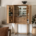 A light natural wood bar cabinet with open doors, styled in a home interior with wine bottles, glassware, and dishes inside. The cabinet features two drawers, glass shelves, and a mirrored back panel, set against a white wall with a staircase in the background.