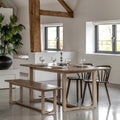 A rustic wooden dining table set in a bright, modern room with exposed beams, a matching wooden bench, and two dark wood chairs. The table is styled with wine glasses, plates, and a candle.