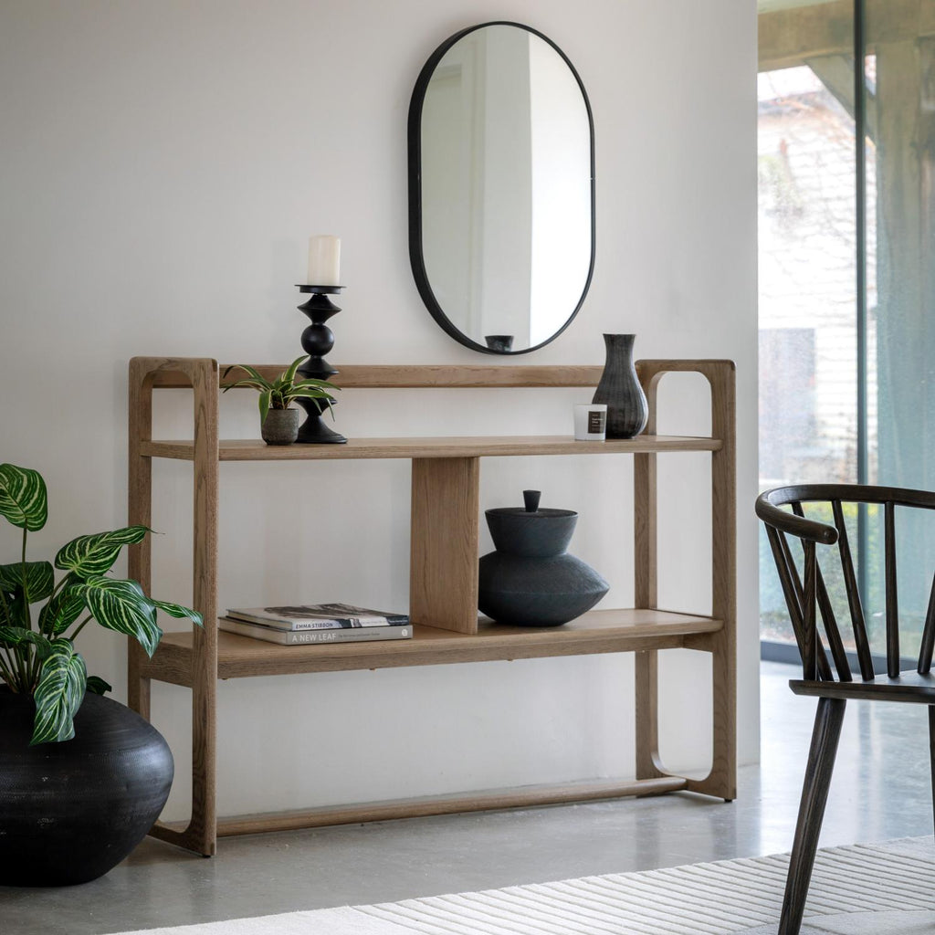 A light natural wood three-tier console shelf styled with black ceramic vases, a candle holder, books, and a small plant, set against a white wall with an oval mirror and a wooden chair nearby.