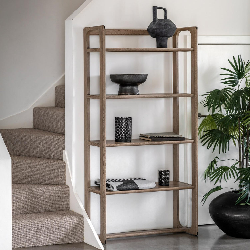 A tall, five-tier wooden shelving unit with a weathered brown finish, styled with home decor items next to a staircase and a potted plant.