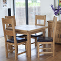 A 5-piece oak dining table set with four wooden chairs featuring black leather seat cushions, shown in a bright dining room setting.