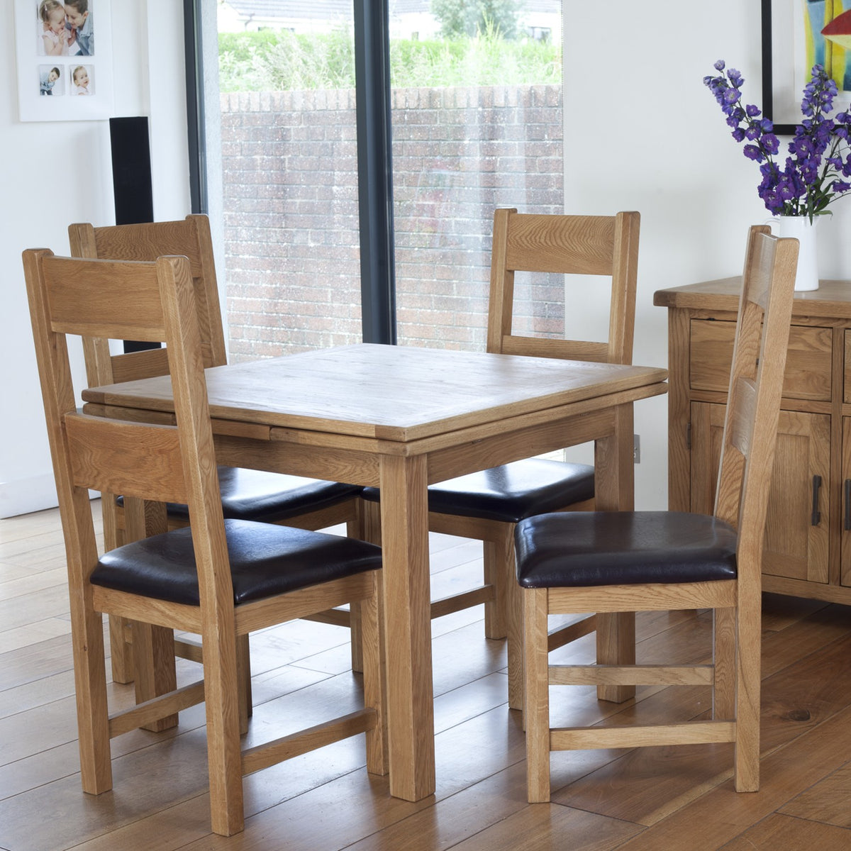 A 5-piece oak dining table set with four wooden chairs featuring black leather seat cushions, shown in a bright dining room setting.