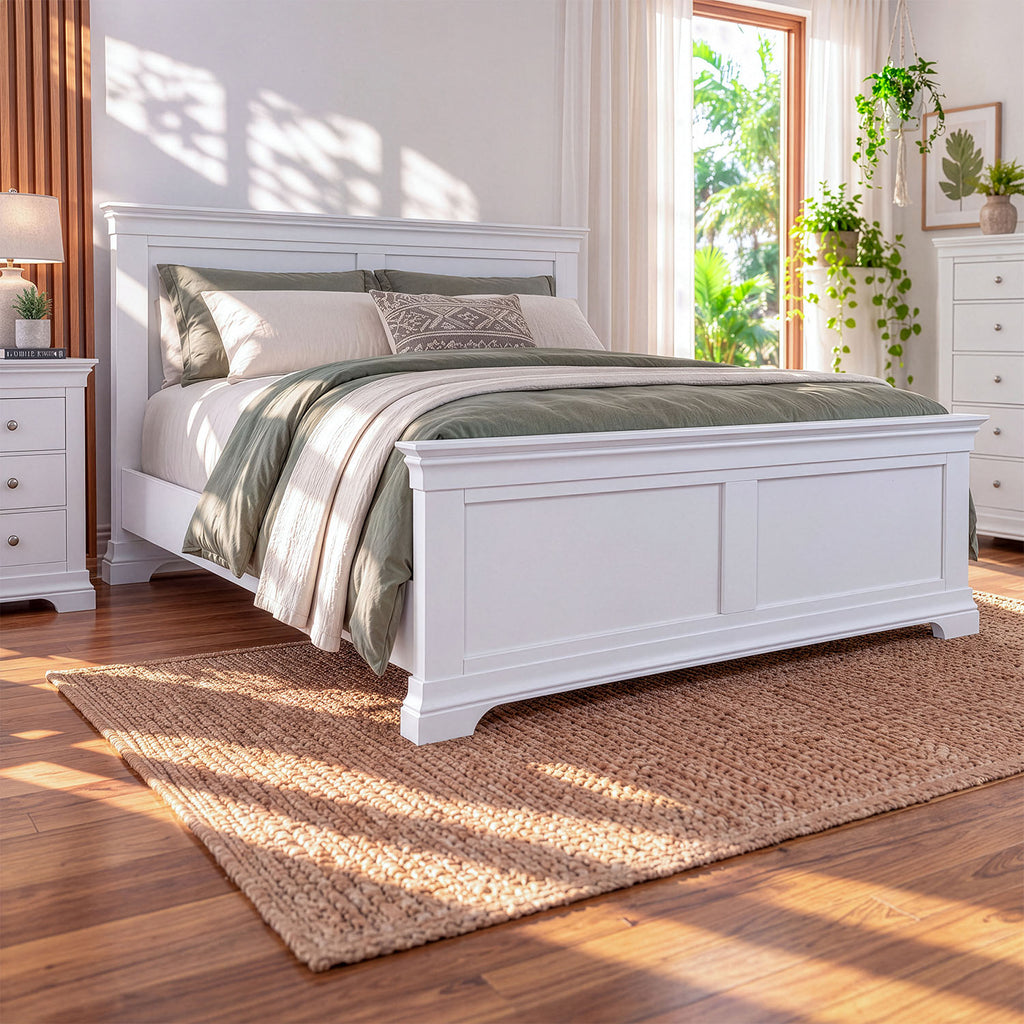 A white wooden bed frame with a panel headboard and footboard, styled with sage green and cream bedding, set in a bright bedroom with hardwood floors, a woven rug, and a window.