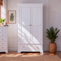 A tall white double-door wardrobe with a single drawer beneath, shown in a bright room with wood flooring, a woven rug, potted plants, and soft sunlight streaming across the front.