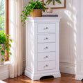 A white five-drawer chest of drawers with small silver knobs, styled in a bright room next to a window with sheer curtains and potted green plants.