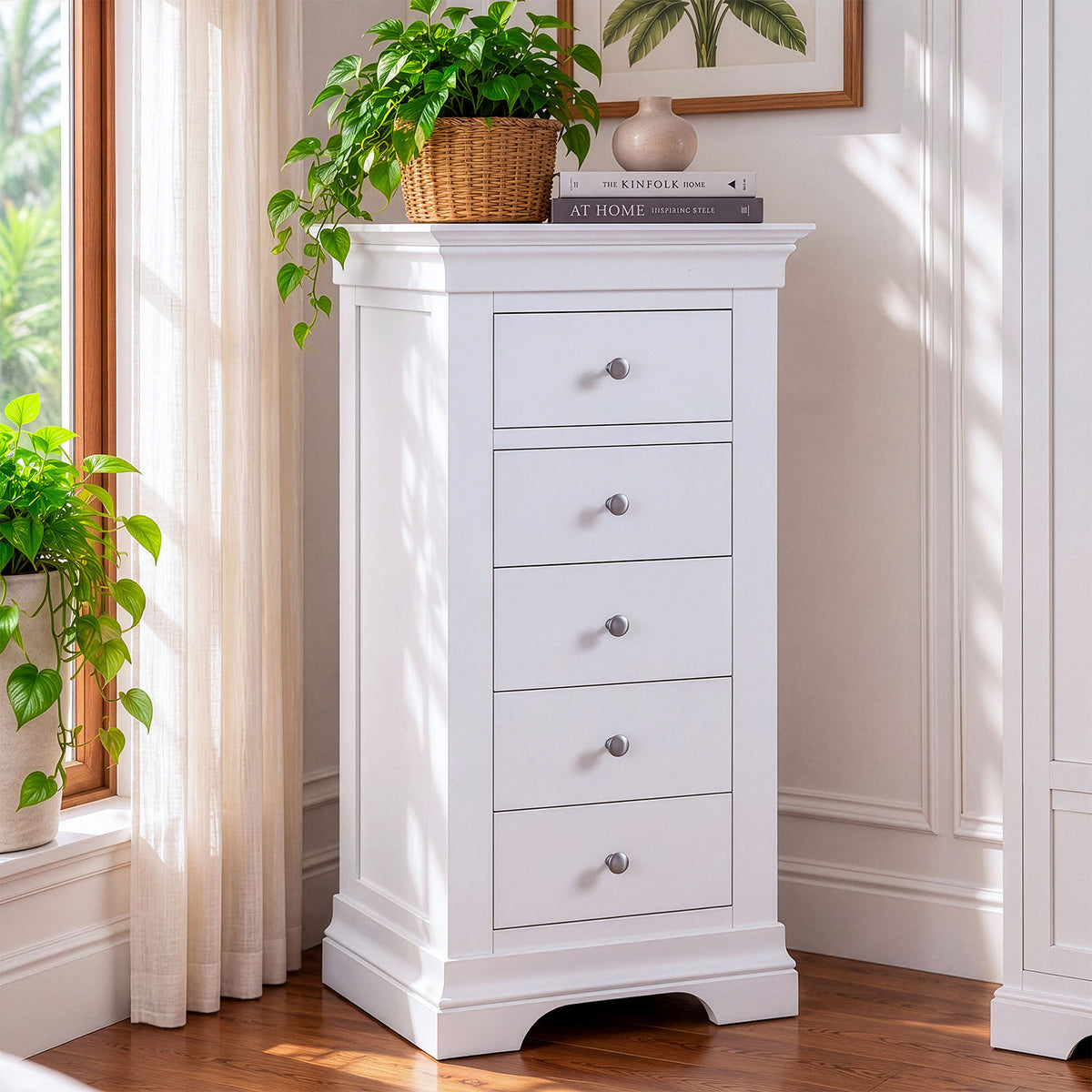 A white five-drawer chest of drawers with small silver knobs, styled in a bright room next to a window with sheer curtains and potted green plants.