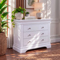 A white three-drawer dresser in a styled room setting, decorated with a potted plant, books, and a ceramic table lamp. The dresser features three drawers with round silver knobs and a curved apron detail at the bottom.