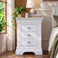 A white three-drawer nightstand in a styled bedroom setting, decorated with a potted green plant, a ceramic table lamp, and stacked books.