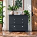 A dark charcoal gray three-drawer dresser styled in a bright, modern room with wooden floors, potted plants, and a framed leaf print on the wall.
