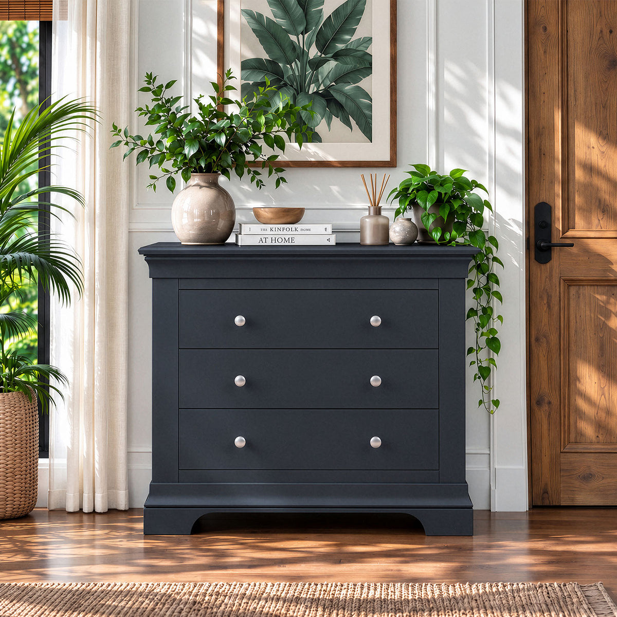 A dark charcoal gray three-drawer dresser styled in a bright, modern room with wooden floors, potted plants, and a framed leaf print on the wall.