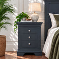 A dark navy nightstand with three drawers and silver knobs, styled in a bedroom next to a bed with white linens, a table lamp, a small potted plant, and a large green houseplant in a wicker basket.