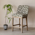 A wooden bar stool with a cream upholstered seat and backrest featuring an olive green abstract pattern, shown next to a potted fern against a neutral beige wall.