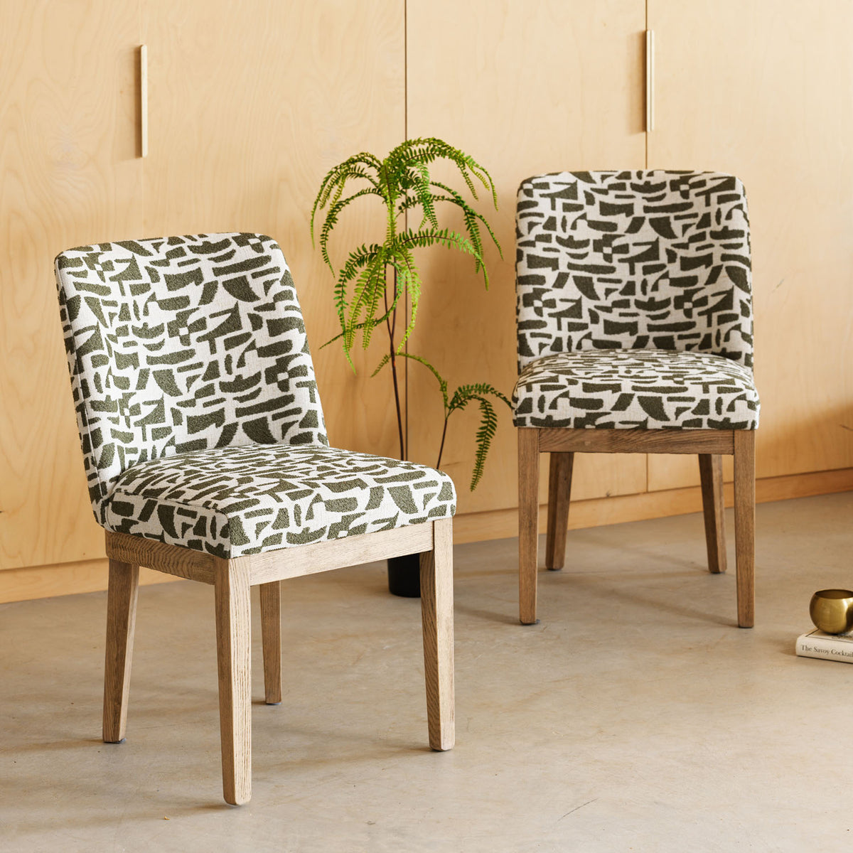 Two upholstered dining chairs with a black and white abstract brushstroke pattern and light natural wood legs, set in a room with a light wood paneled wall and concrete floor.