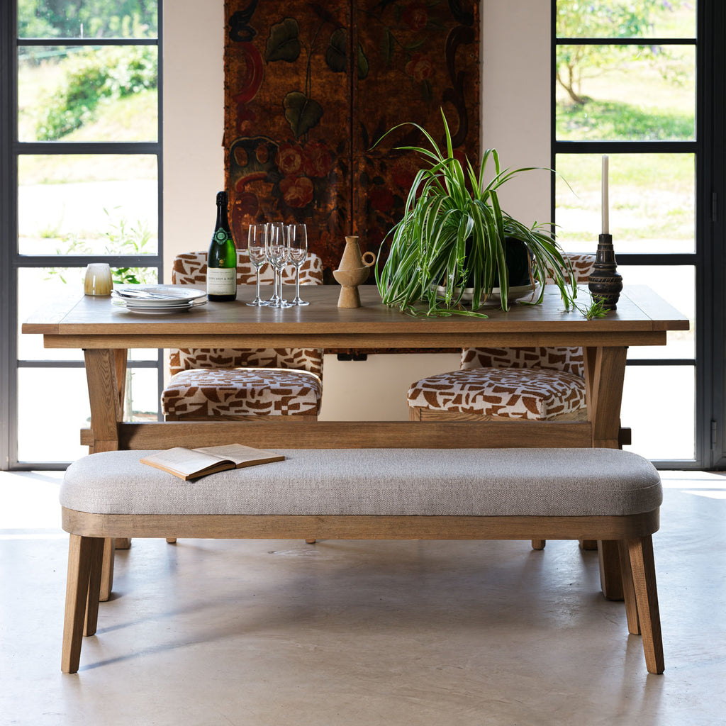 A rustic wooden dining table set in a bright room with tall black-framed windows, a matching upholstered bench, patterned chairs, a potted green plant, and dining glassware.