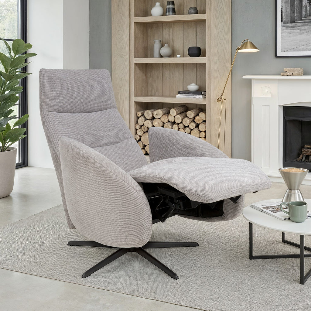 A light gray upholstered recliner chair with a black swivel base, shown in a modern living room setting with a fireplace, shelving, and a small side table.