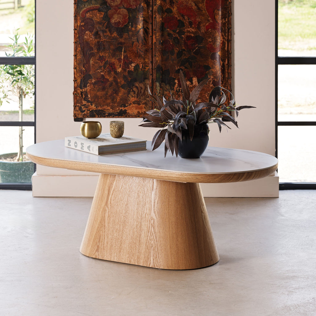 An oval coffee table with a light oak wood pedestal base and a white marble top, styled in a modern interior with a potted plant and books on top.