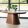 A modern oval coffee table with a dark walnut wood pedestal base and a light gray stone top, styled with a potted green plant and a candle in front of a large black-framed window.