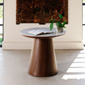 A round wooden accent table with a tapered base and a light gray stone top, styled in a bright room with an open book and a small potted plant on the surface.