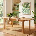 A light natural wood dining table set with plates, folded napkins, and a large floral centerpiece in a vase, styled in a bright room with green walls, plants, shelves, and a woven pendant light.