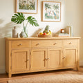 A light natural wood sideboard cabinet with four doors and three drawers, styled in a warm room setting with decorative vases, a fruit bowl, books, and framed tropical wall art.