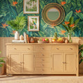A tropical-themed interior scene featuring a light wood sideboard cabinet against a bold green palm-leaf wallpaper background. The cabinet is styled with books, vases, fruit baskets, and plants, with framed tropical art and a round woven mirror above.