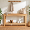 A light natural wood entryway bench with an open lower shelf, styled with a cream throw blanket, a beige tote bag, a pair of brown ankle boots, and white sneakers. The bench is set against a warm off-white wall with a wooden coat rack and shadow details, flanked by green plants.