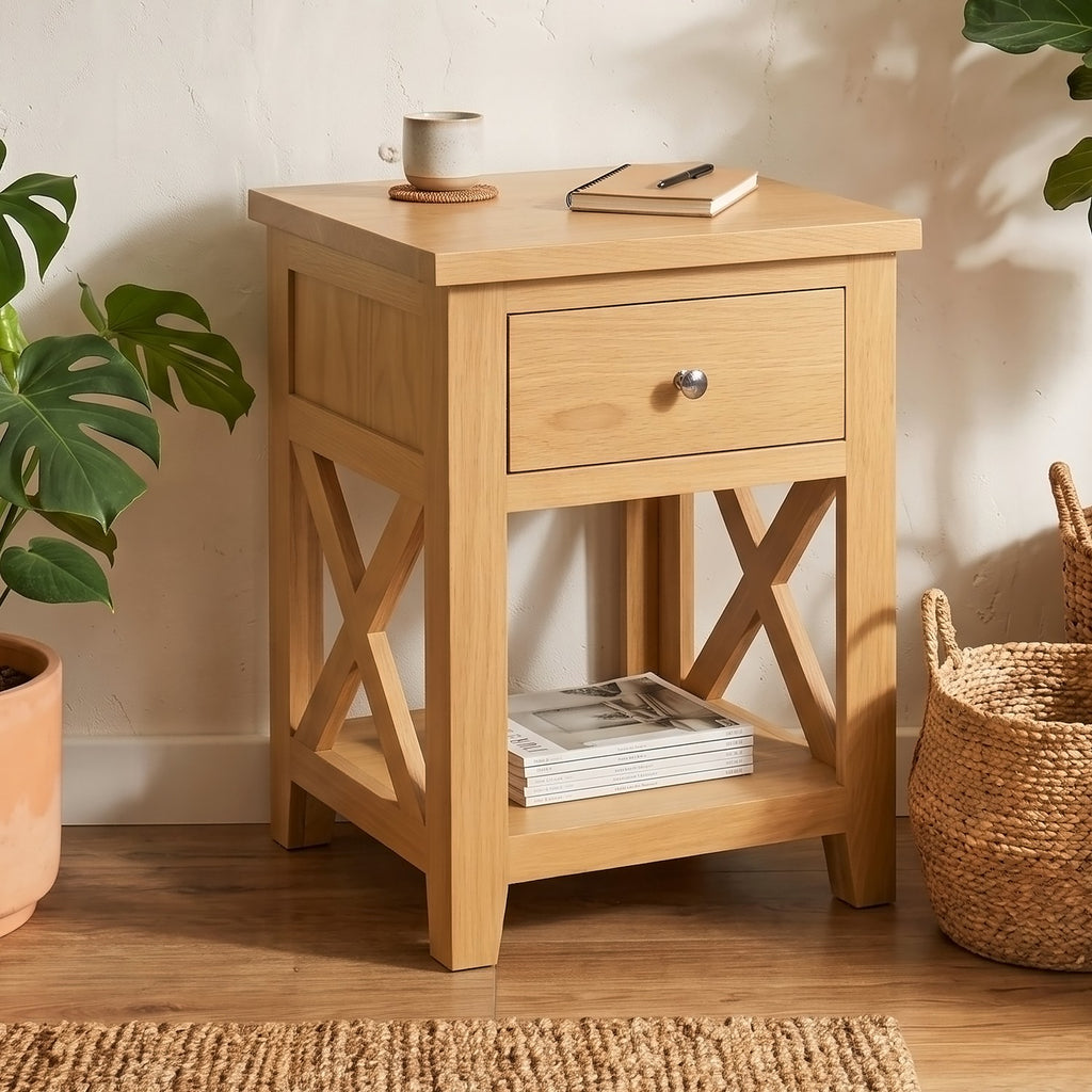 A light natural wood bedside table with one drawer and an open lower shelf, styled in a room setting with a mug, notebook, books, plants, and woven baskets.
